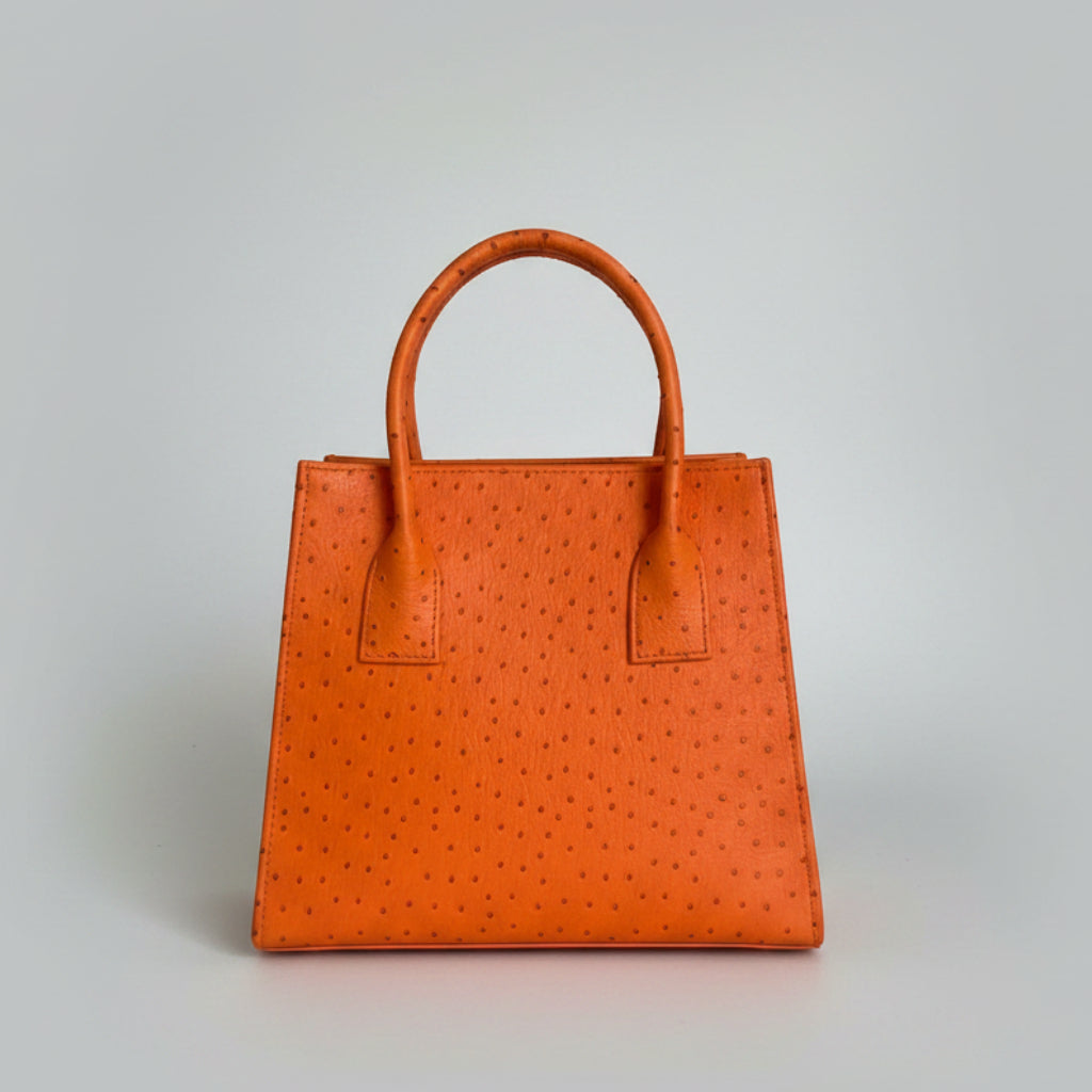 An orange ostrich-textured leather handbag with double top handles, displayed on a marble surface with a decorative background.