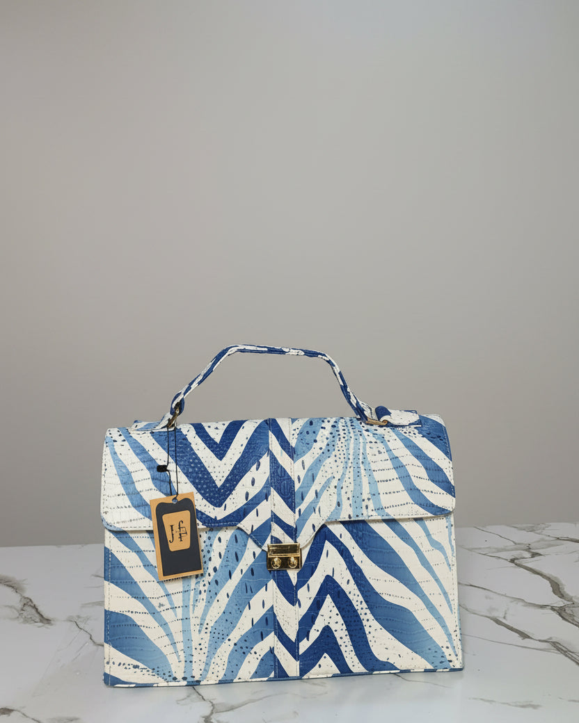 A structured handbag with a blue and white chevron pattern, featuring a top handle, gold-tone hardware, and a hanging brand tag, displayed on a marble surface in front of a branded wall sign.