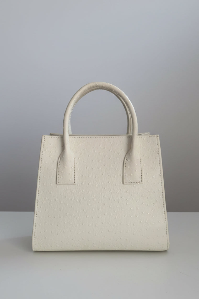 A cream-colored ostrich leather handbag with double top handles, displayed on a white marble surface with a neutral indoor background.
