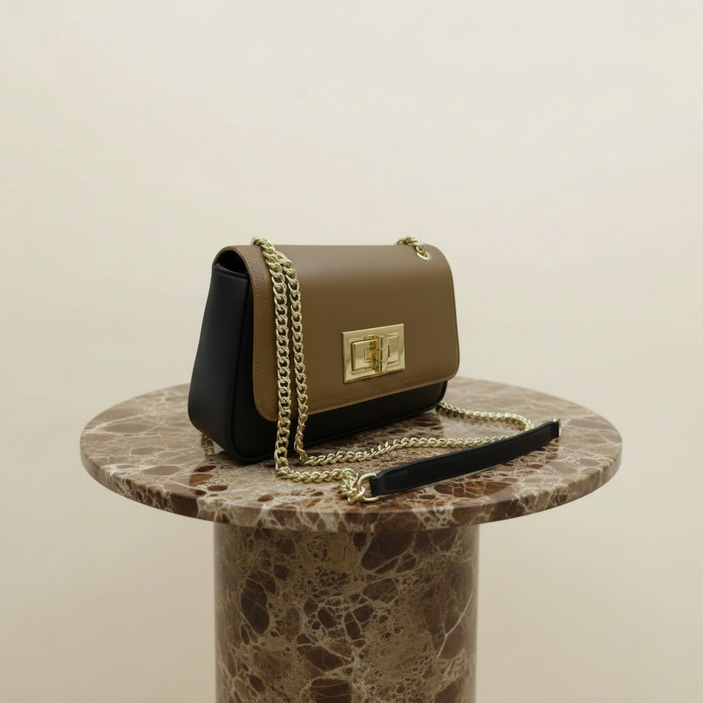 A structured shoulder bag with a two-tone design, featuring a tan leather flap and a black leather body, accented with a gold-tone chain strap and a white protective wrap on the strap. The bag is displayed on a wooden stand against a modern wall with a yellow wave-like light.
