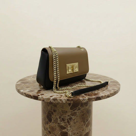 A structured shoulder bag with a two-tone design, featuring a tan leather flap and a black leather body, accented with a gold-tone chain strap and a white protective wrap on the strap. The bag is displayed on a wooden stand against a modern wall with a yellow wave-like light.