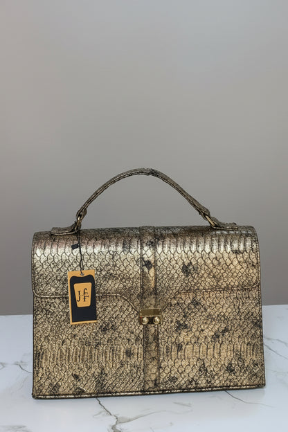 A structured snakeskin handbag in metallic gold and brown tones, featuring a top handle and a small hanging brand tag, displayed on a marble surface with a branded wall sign behind it.