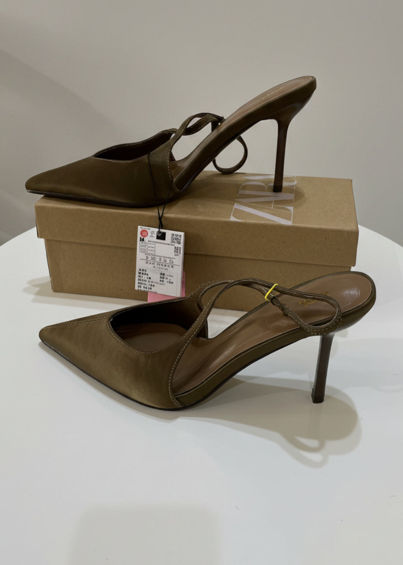 A pair of taupe-brown pointed-toe slingback high heels resting on a brown shoebox on a white table, with a decorative vase and pampas grass in the background.