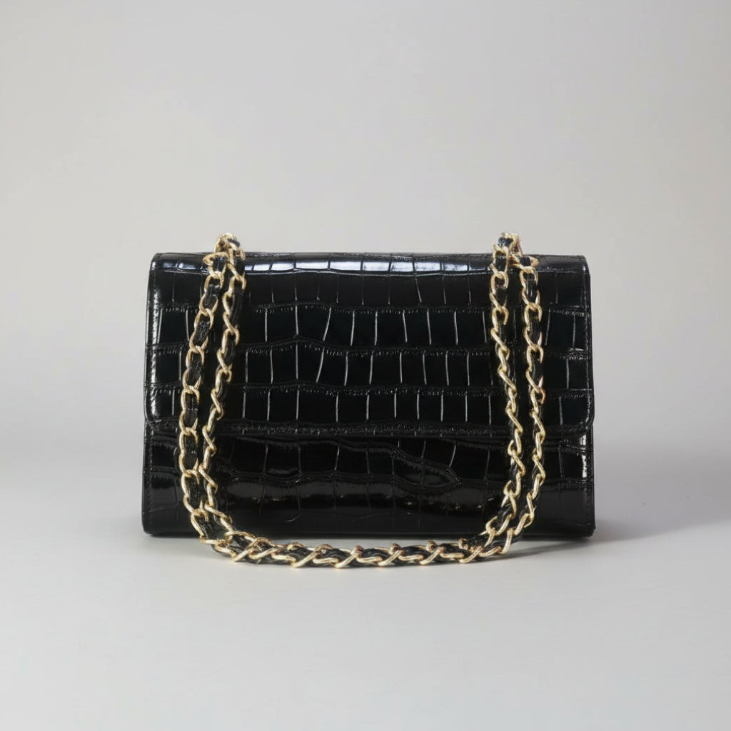 A black crocodile-embossed leather handbag with a gold-tone chain strap, resting on a light wooden surface with a blurred green plant in the background.