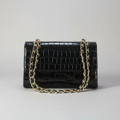 A black crocodile-embossed leather handbag with a gold-tone chain strap, resting on a light wooden surface with a blurred green plant in the background.