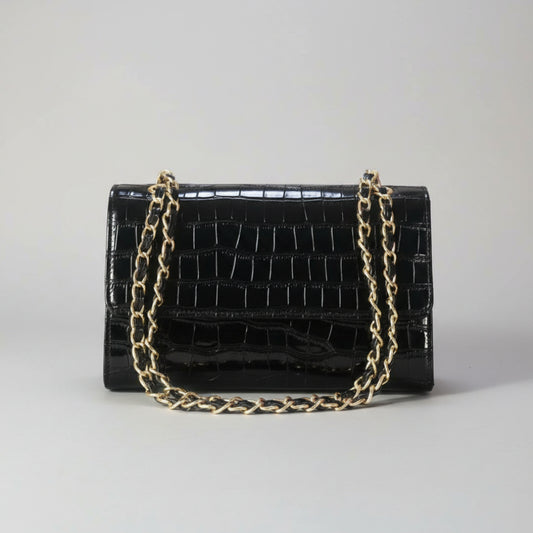 A black crocodile-embossed leather handbag with a gold-tone chain strap, resting on a light wooden surface with a blurred green plant in the background.