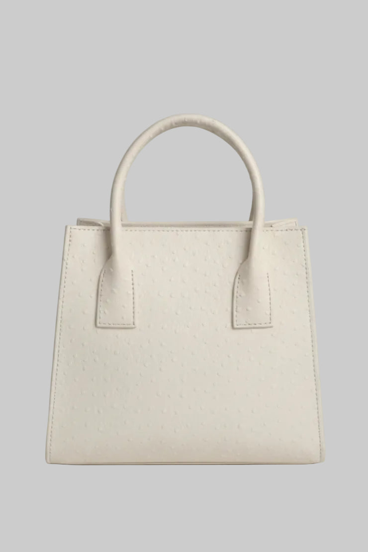 A cream-colored ostrich leather handbag with double top handles, displayed on a white marble surface with a neutral indoor background.