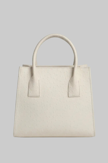 A cream-colored ostrich leather handbag with double top handles, displayed on a white marble surface with a neutral indoor background.