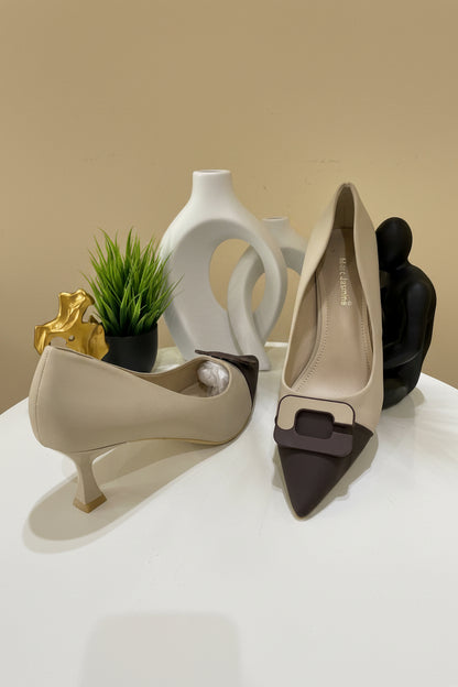 A pair of women's pointed-toe pumps in beige and dark brown, styled on a white tabletop with decorative vases, a small plant, and dried grass in the background.