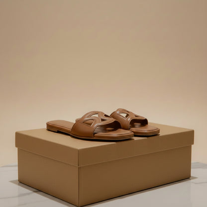 A pair of tan leather slide sandals with a decorative cutout logo on the straps, displayed on a cardboard shoe box on a white surface with a decorative vase in the background.