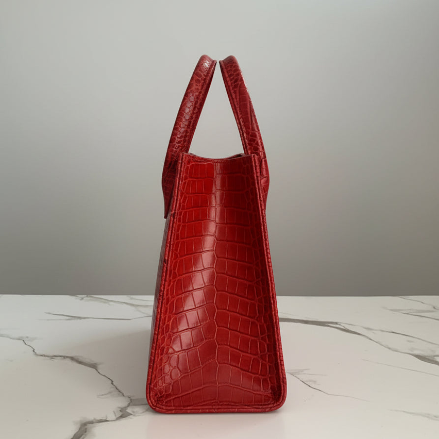 A bright red python-textured handbag with double top handles and a gold-tone ring detail, standing on a white marble surface against a decorative background.