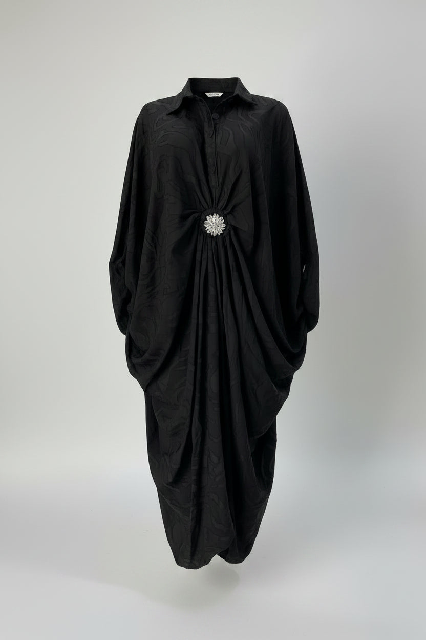 A black long-sleeve collared dress displayed on a mannequin in a boutique setting. The dress features a gathered, ruched design at the waist with a prominent silver rhinestone brooch at the center front.