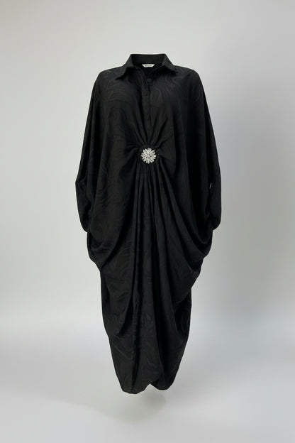 A black long-sleeve collared dress displayed on a mannequin in a boutique setting. The dress features a gathered, ruched design at the waist with a prominent silver rhinestone brooch at the center front.