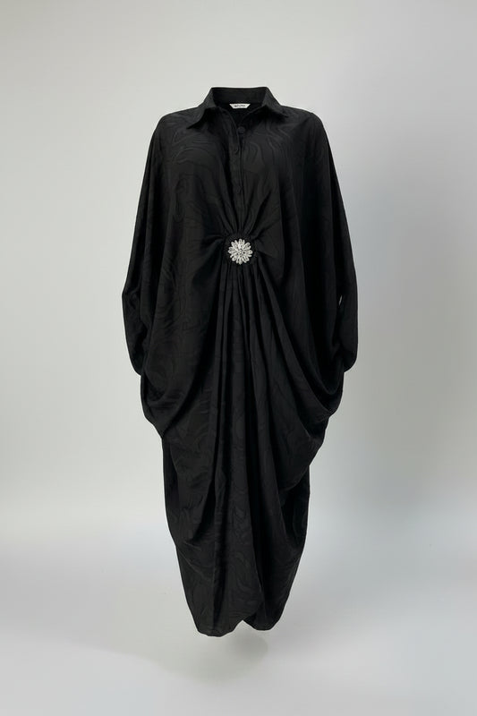 A black long-sleeve collared dress displayed on a mannequin in a boutique setting. The dress features a gathered, ruched design at the waist with a prominent silver rhinestone brooch at the center front.